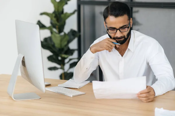 Hombre indio barbudo serio concentrado, hombre de negocios, con un atuendo formal, sentado en una mesa en la oficina, estudiando documentos, analizando un informe y cartas financieras — Foto de Stock