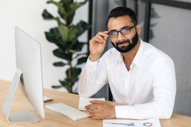 Pozitif başarılı Hintli bir iş adamı, menajer ya da bilişim uzmanı portresi, beyaz gömlekli, gözlüklü, ofisinde masasında oturuyor, kameraya bakıyor, hoş ve dost canlısı görünüyor.
