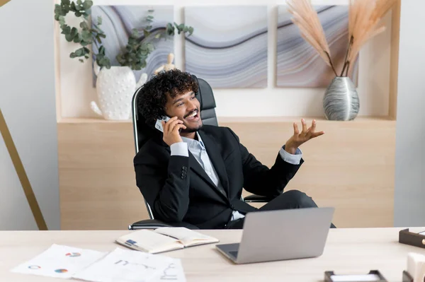 Ocupado bem sucedido empresário indiano, especialista em TI, proprietário da empresa, sentado na mesa de trabalho em casa, tendo uma conversa de telefone celular de prazer durante o tempo de trabalho, sorriso amigável, desvios — Fotografia de Stock