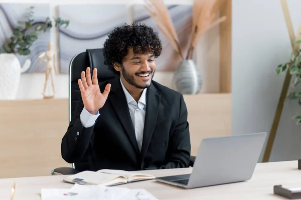 Онлайн видео связь. Позитивный успешный индийский бизнесмен в официальном костюме, сидит за столом в офисе, использует ноутбук для общения через видео-звонок, разговаривает с коллегой или другом, улыбается — стоковое фото
