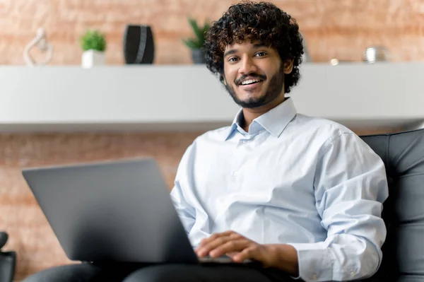 Indiase positieve zelfverzekerde krullende jonge volwassen man, freelancer of manager in shirt, zittend op een fauteuil met laptop kijken naar de camera en glimlachend vriendelijk — Stockfoto