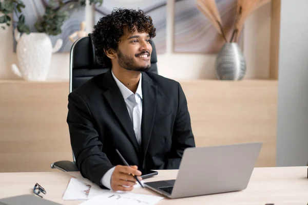 Indisk positiv forretningsmann, i formelle klær, sittende ved bordet i embetet, mens han jobber i en bærbar datamaskin, ser positivt på, tenker på en oppstart eller drømmer om en fremtidig ferie stockbilde