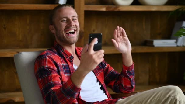 Διαδικτυακή επικοινωνία βίντεο. Θετικός Αμερικανός νεαρός άνδρας, κάθεται σε πολυθρόνα στο σπίτι, χρησιμοποιεί κινητό τηλέφωνο, μιλάει σε βιντεοκλήση με φίλους ή συγγενείς, συνομιλεί σε κοινωνικά δίκτυα, χαμογελάει — Αρχείο Βίντεο