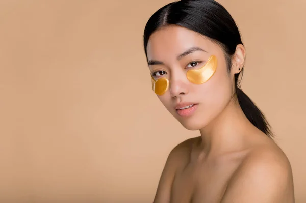 Gold eye patches for puffiness, wrinkles and dark circles, skincare concept. Pretty young Asian brunette woman with perfect skin applied gold collagen eye patches,standing on isolated beige background