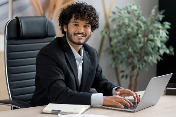 Retrato de un joven empresario indio de pelo rizado carismático positivo, vestido de negocios, sentado en su escritorio en la oficina, mirando a la cámara con una sonrisa amistosa —  Fotos de Stock