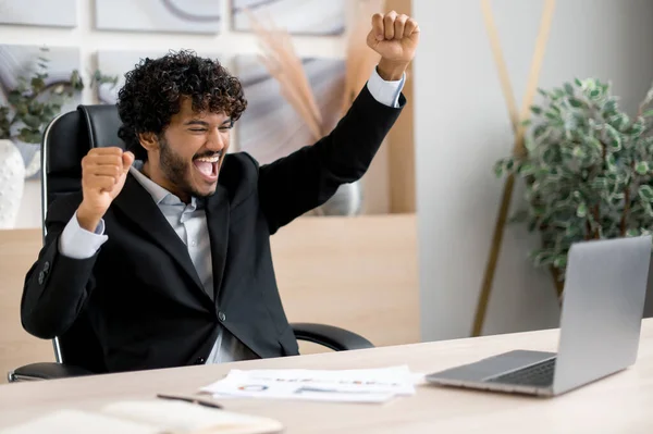 Succesvolle blijde Indiase zakenman, manager, aandelenhandelaar, zit aan een tafel in het kantoor, schreeuwt en verheugt zich over succes, overwinning of goede winst, kijkt naar een laptop en gebaren met zijn handen — Stockfoto
