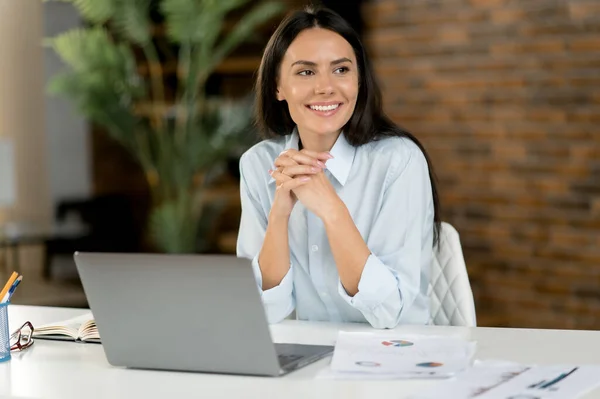 Atraktivní příjemná kavkazská brunetka podnikatelka, mentorka, kancelářská pracovnice, finanční poradkyně, sedící za stolem v kanceláři, dívající se stranou, přátelsky se usmívající, si dává pauzu od práce — Stock fotografie