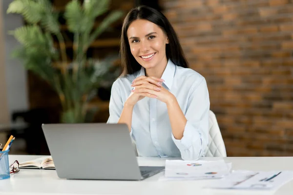 Portret van een blanke brunette vrouw. Succesvolle vrouwelijke kantoormedewerker, topmanager, bedrijfsadviseur, draagt formeel shirt, zit op haar werkplek met laptop, kijkt naar de camera, glimlachend vriendelijk — Stockfoto