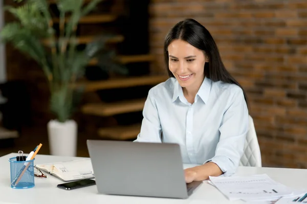 Positieve vrij Kaukasische brunette vrouw, top manager, ondernemer, secretaresse, makelaar, werken met laptop, zit op haar werkplek in het kantoor, met behulp van netwerk, glimlachend vriendelijk — Stockfoto