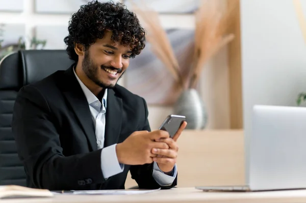 스마트폰으로 온라인 채팅. 만족 한 인도 남자, 최고 관리자, 비즈니스 소유자는 책상에 앉아 일하면서 핸드폰을 사용하고 인터넷을 검색하고 이메일을 받고 친구들 과 문자를 보내고 미소를 짓습니다. — 스톡 사진