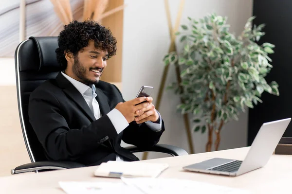 Comunicación remota usando un smartphone. indio positivo hombre sentado en su escritorio en la oficina, mensajes de texto en línea su gadget, respuestas de correo electrónico, navega por Internet, sonriendo amigable —  Fotos de Stock