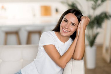 Güzel, beyaz saçlı, uzun saçlı, yetişkin bir kadın. Günlük beyaz tişört giyiyor. Oturma odasında kanepede oturuyor.