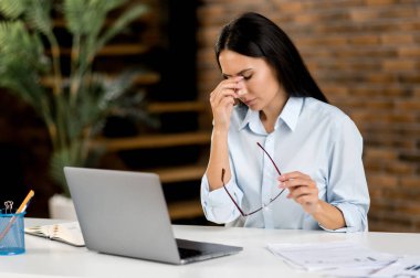 Baş ağrısı ve migren. Yorgun, Kafkasyalı genç bir kadın, üst düzey yönetici, ofis çalışanı, çalışmaktan yorulmuş, stres altında, dinlenmeye ihtiyacı var, burnunun köprüsüne masaj yapıyor, gözlerini kapatıyor.