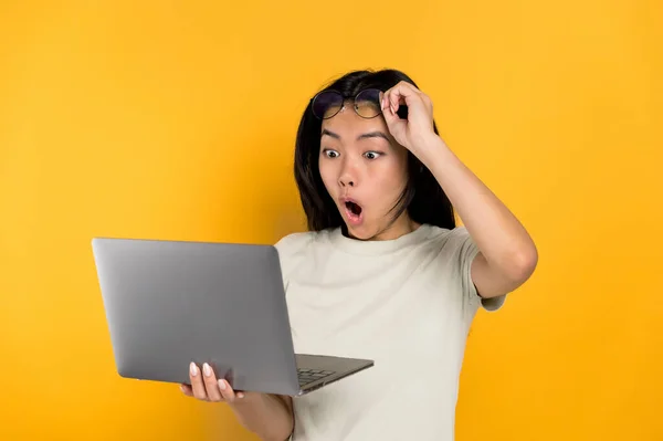 Здивований шокований японець молода брюнетка, тримаючи відкритий ноутбук в руці, виглядає здивованим, знімає окуляри, відкриваючи рот, стоячи на ізольованому помаранчевому фоні в повсякденній футболці — стокове фото