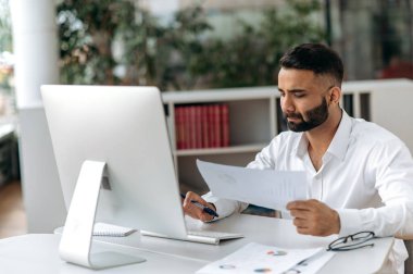 Amaçlı genç Hintli işadamı, menajer ya da sakallı bir ofis çalışanı, beyaz gömlek giyiyor, masasında oturuyor, bilgisayar kullanıyor, belgeler ve şemalar üzerinde çalışıyor, analitik