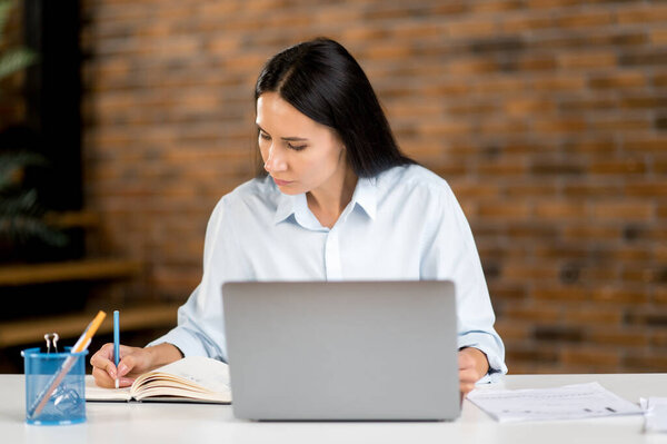 Serious attractive successful smart focused young caucasian brunette business woman, manager or freelancer, sitting at table in the office, in stylish clothes, using laptop, taking notes