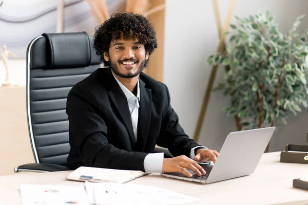 Portret van jonge volwassen knappe vrolijke zelfverzekerde succesvolle Indiase zakenman, CEO of manager met baard, in zakelijke kleding, zittend aan zijn bureau in het kantoor met een vriendelijke glimlach — Stockfoto
