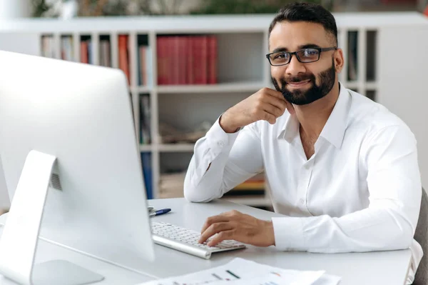 Retrato exitoso inteligente feliz guapo empresario indio, freelancer o gerente con barba, con camisa blanca, sentado en la mesa con el ordenador en la oficina, mirando a la cámara y sonriendo amigable — Foto de Stock