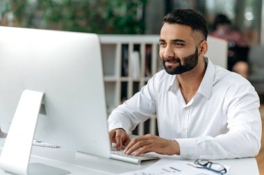 Odaklanmış zeki, başarılı bir Hintli işadamı, finans yöneticisi ofiste oturuyor, tişört giyiyor, bilgisayar kullanıyor, iş arkadaşlarıyla mesajlaşıyor, notlar alıyor, finansal bir strateji geliştiriyor.