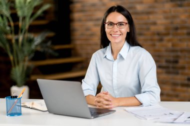 Kendine güvenen, zeki, güzel, beyaz tenli, iş kadını CEO ya da yönetici portresi. Ofiste oturuyor, dizüstü bilgisayar kullanıyor, resmi kıyafetler giyiyor, kameraya bakıyor, gülümsüyor.