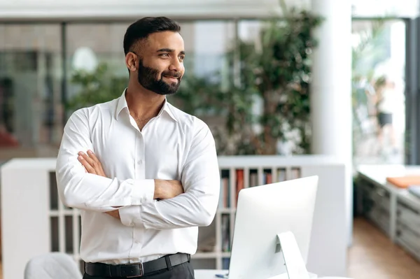 Retrato bonito confiante, inteligente jovem adulto indiano empresário, freelancer ou gerente de topo com barba em camisa elegante branco de pé perto da mesa no escritório olhar para o lado sorrindo no fundo borrado — Fotografia de Stock
