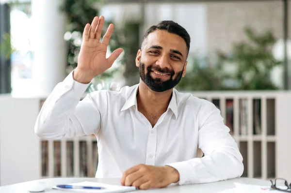 Retrato confiado alegre joven adulto empresario indio, gerente con barba, en camisa blanca, sentado en la mesa en la oficina, comunicando colegas a través de la comunicación de vídeo, agitando la mano, sonriendo — Foto de Stock