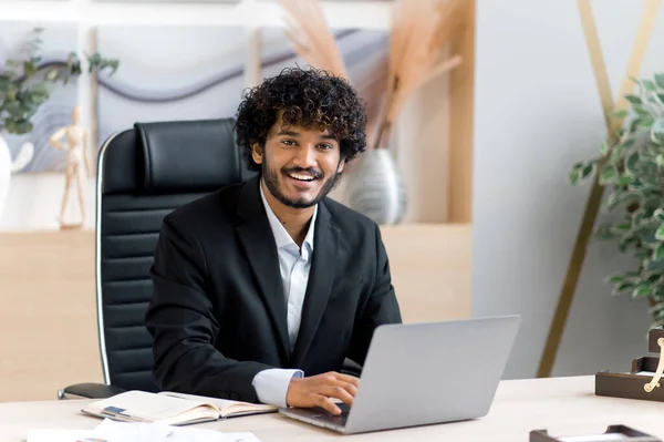 Joyful jovem adulto bem sucedido atraente encaracolado indiana top manager, CEO ou empresário em um terno elegante senta-se a uma mesa em um escritório moderno, usa um laptop, olha para a câmera, sorri — Fotografia de Stock