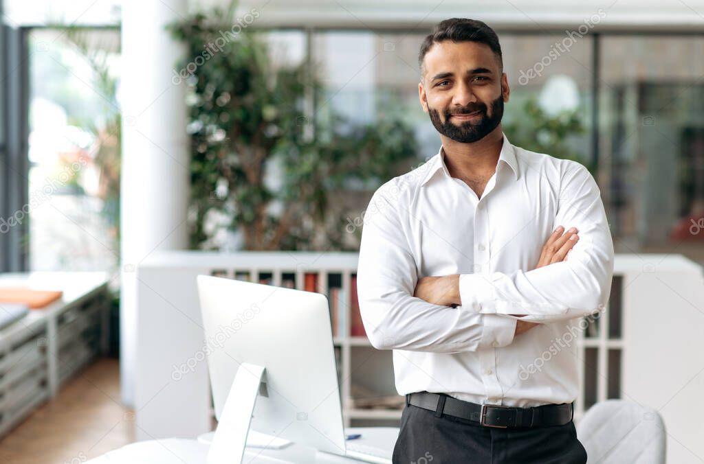 Portrait of attractive, confident, smart, influential handsome Indian business leader, CEO, top manager, IT specialist, in a white shirt, stand near the table in the office, looks at the camera smiles