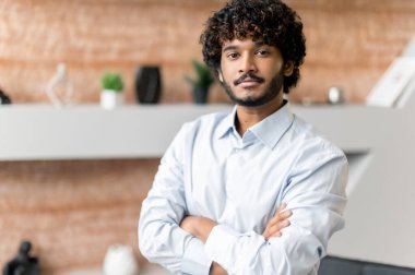 Çekici, modern, kıvırcık saçlı, sakallı, ofis çalışanı, serbest çalışan, üst düzey yönetici, kollarını kavuşturmuş vaziyette ofiste kameraya bakan,