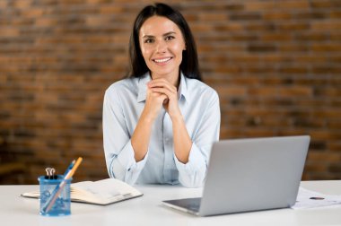 Şirin, kendine güvenen, genç, başarılı, Kafkasyalı ofis çalışanı, iş kadını, resmi tişört giymiş yönetici iş yerinde dizüstü bilgisayarla oturmuş gülümsüyor.