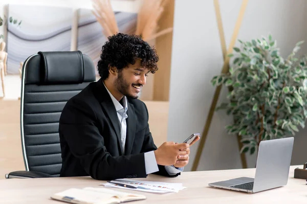 Tevreden jonge volwassen indiaan, topmanager, zakenman aan tafel, smartphone gebruiken tijdens het werk, surfen op internet of nieuws, e-mail beantwoorden, sms 'en met vrienden, glimlachen — Stockfoto