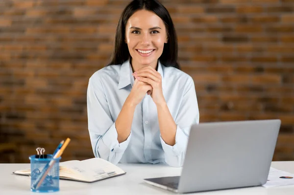 Portret jong volwassene succesvolle mooie blanke zakenvrouw, kantoormedewerker, manager, business consultant, gekleed in formeel shirt, zitten op haar werkplek met laptop, kijken naar camera, glimlach — Stockfoto