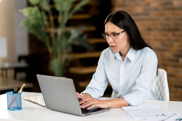 Serious puzzled young adult caucasian brunette business woman, ceo or top manager wearing glasses sitting in office, using laptop, analyzing financial market, preparing business strategy or report — Stock Photo, Image