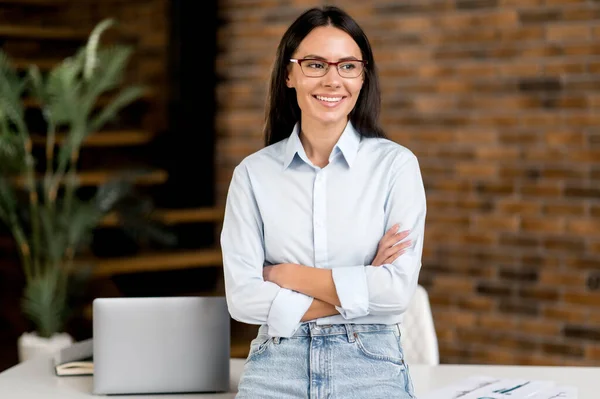 Portret succesvolle schattige zelfverzekerde mooie jonge volwassen blanke zakenvrouw of makelaar staan in de buurt van het werk bureau in kantoor gekleed in stijlvolle kleding, armen gekruist, kijken naar opzij, glimlachen — Stockfoto