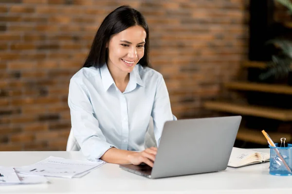 Blank aantrekkelijk zelfverzekerd jong volwassen zakenvrouw, top manager of makelaar, brunette, zittend in modern kantoor, in stijlvolle kleding, met behulp van laptop voor het ontwikkelen van nieuw project, glimlachen — Stockfoto