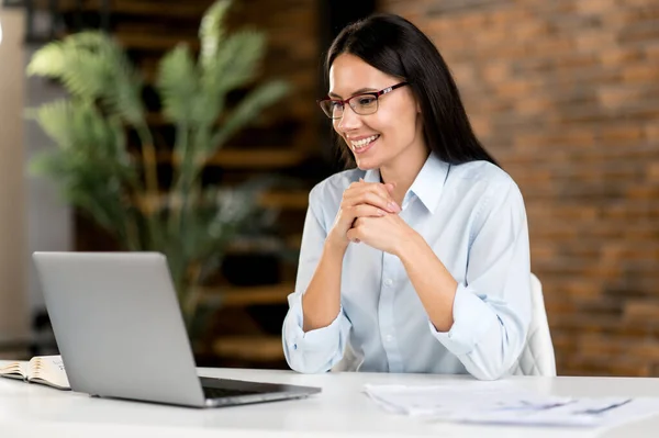 Przyjazna, ładna, odnosząca sukcesy biała brunetka bizneswoman, broker, corporate manager, używająca laptopa, używa laptopa siedząc przy biurku, planując projekt, gestykulując, uśmiechając się szczęśliwie — Zdjęcie stockowe