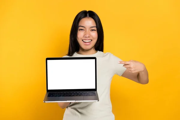 Spännande kinesisk positiv ung kvinna i casual t-shirt hålla öppen bärbar dator med tom vit skärm och pekar ett finger åt honom, står på isolerad orange bakgrund, tittar på kamera, mockup, kopiera utrymme — Stockfoto