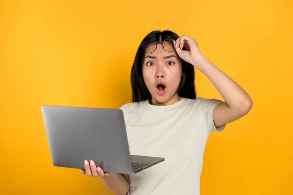 Вражена шокована китайська молода брюнетка, що тримає відкритий ноутбук в руці, виглядає здивованою в камері, знімає окуляри, відкриваючи рот, стоячи на ізольованому помаранчевому тлі в випадковій футболці — стокове фото