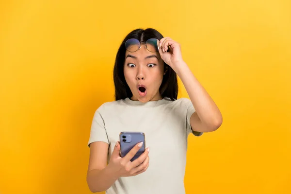 Erstaunt schaut die junge brünette Japanerin, die ein Handy in der Hand hält, überrascht drein, nimmt ihre Brille ab und öffnet den Mund, steht auf orangefarbenem Hintergrund in lässigem T-Shirt — Stockfoto