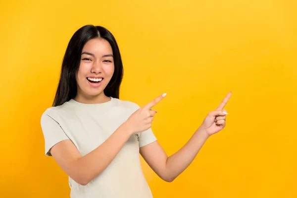 Verwirrt beeindruckt brünette junge Chinesin aus dem Millennium, zeigt mit den Händen auf den leeren Raum, blickt in die Kamera, lächelt freundlich, steht auf isoliertem Hintergrund in lässigem Hemd — Stockfoto