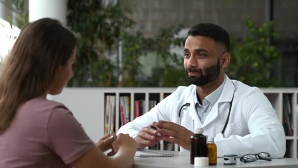 Overleg met een arts in een kliniek. Indiase huisarts in een medisch uniform, adviseert de patiënt, schrijft behandeling voor, luistert naar klachten, geeft advies, toont geneesmiddelen — Stockvideo