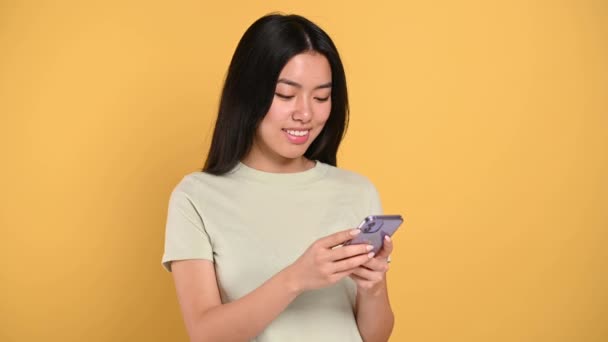 Excited joyful, pretty Asian long-haired brunette girl, in casual t-shirt, holds a smartphone in her hands, chatting with friends in social media, texting, stands on isolated orange background, smile — Stockvideo