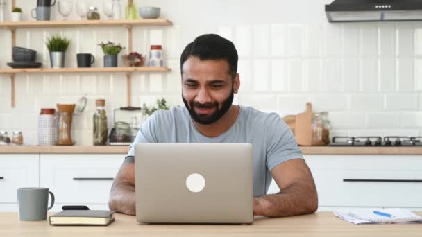 Happy excited lucky Indian man, in casual wear, freelancer, sits at a desk in kitchen, work from home, gesturing with his fists rejoicing in victory, success, good deal, big profit — Stock Video