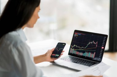 Genç, kendine güvenen kadın yatırımcı, borsacı veya kripto tüccarı, dizüstü bilgisayar ve akıllı telefon kullanarak, kripto sikkeleri alıp satıyor, borsa ve dijital kripto döviz borsalarında ticaret grafiklerini analiz ediyor.