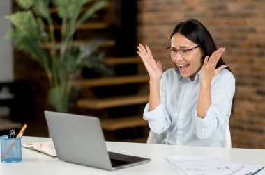 Şaşırmış genç esmer iş kadını, menajer ya da simsar masada oturuyor, dizüstü bilgisayar kullanıyor, haberlerden ya da mesajdan şok geçiriyor, maddi kayıp yaşıyor, şok geçiriyor, çığlık atıyor.