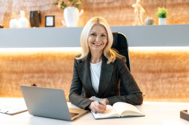 Zarif, orta yaşlı beyaz bir kadının portresi, iş kadını, üst düzey bir yönetici, takım elbiseli, ofiste masada oturuyor, kameraya bakıyor, dostça gülümsüyor.