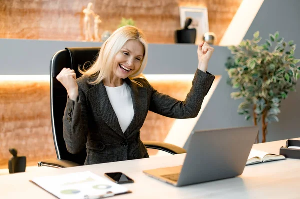 Succesvolle vrolijke opgewonden blanke volwassen zakenman, manager, aandelenhandelaar, zit aan een tafel in het kantoor, schreeuwt en verheugt zich over succes, overwinning of goede winst, kijkt naar een laptop, gebaren handen — Stockfoto