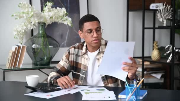 Εστιασμένος σύγχρονος ισπανόφωνος νέος με γυαλιά, υπάλληλος γραφείου ή ελεύθερος επαγγελματίας, κάθεται στο γραφείο εργασίας του, βλέπει γραφήματα, αναλύει εκθέσεις και κέρδη, εργάζεται με έγγραφα στο γραφείο — Αρχείο Βίντεο