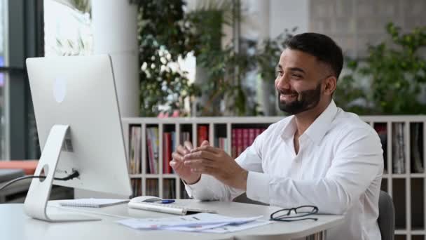 Счастливый дружелюбный, уверенный в себе успешный индийский бизнесмен или генеральный директор с бородой, сидящий в офисе, одетый в белую рубашку, использующий компьютер для личного общения, машущий рукой, улыбающийся — стоковое видео