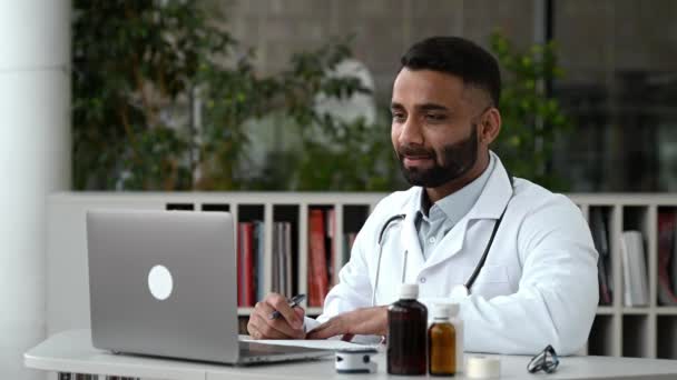 Orgulloso terapeuta médico indio inteligente lleva a cabo una consulta en línea. El médico masculino se comunica con su paciente mediante videollamada utilizando una computadora portátil, prescribe el tratamiento, responde preguntas, hace recomendaciones — Vídeo de stock